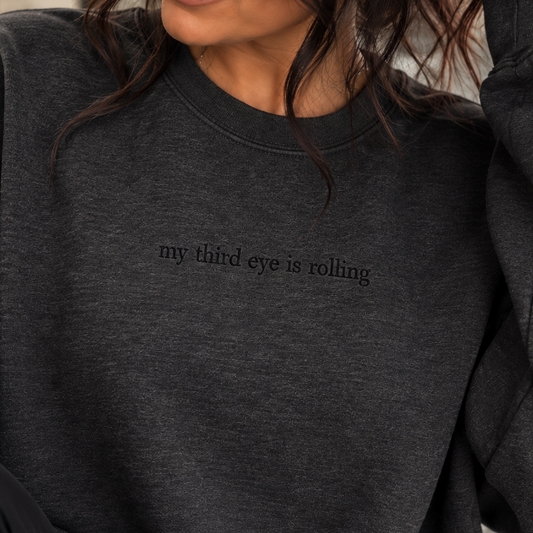 A woman wearing a dark charcoal gray crewneck sweatshirt with the text 'my third eye is rolling' printed in black across the chest, set against a softly blurred indoor background.