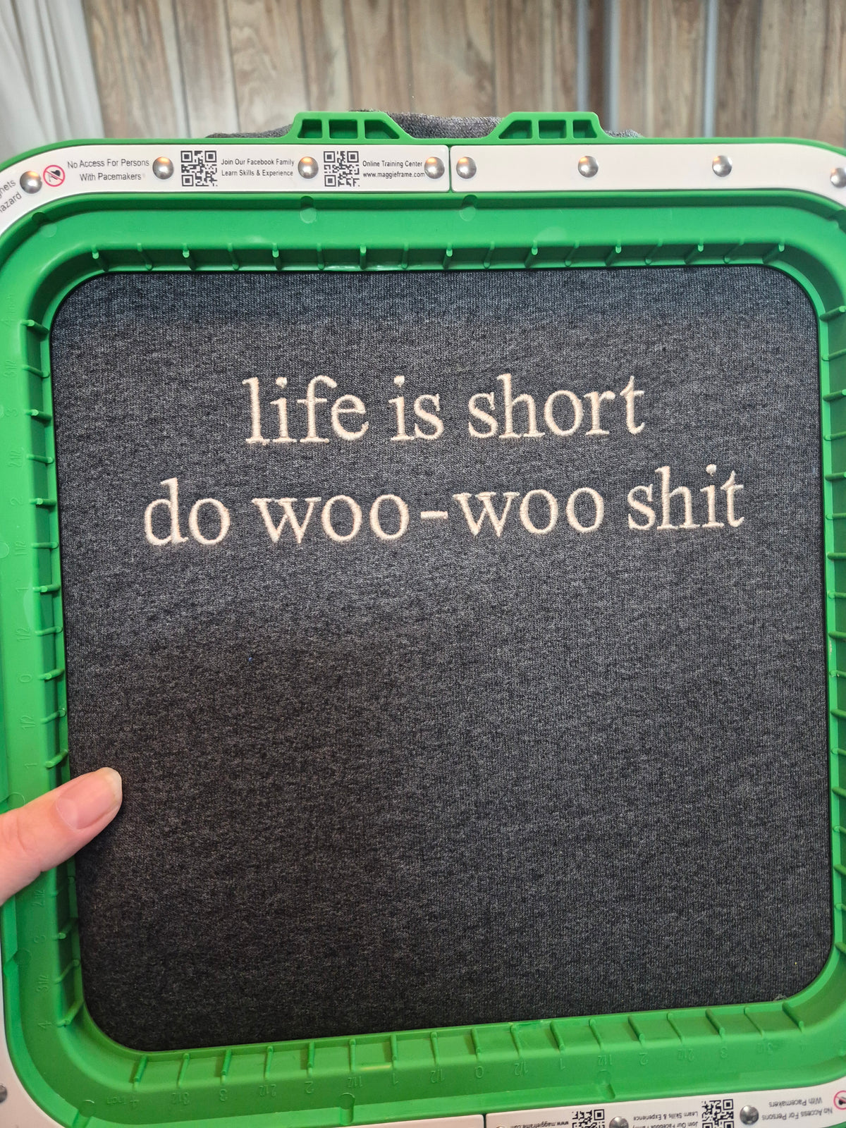 A dark gray felt embroidery hoop cover with the phrase 'life is short do woo-woo shit' embroidered in light beige text, held in a green embroidery machine frame with a finger visible at the bottom left.