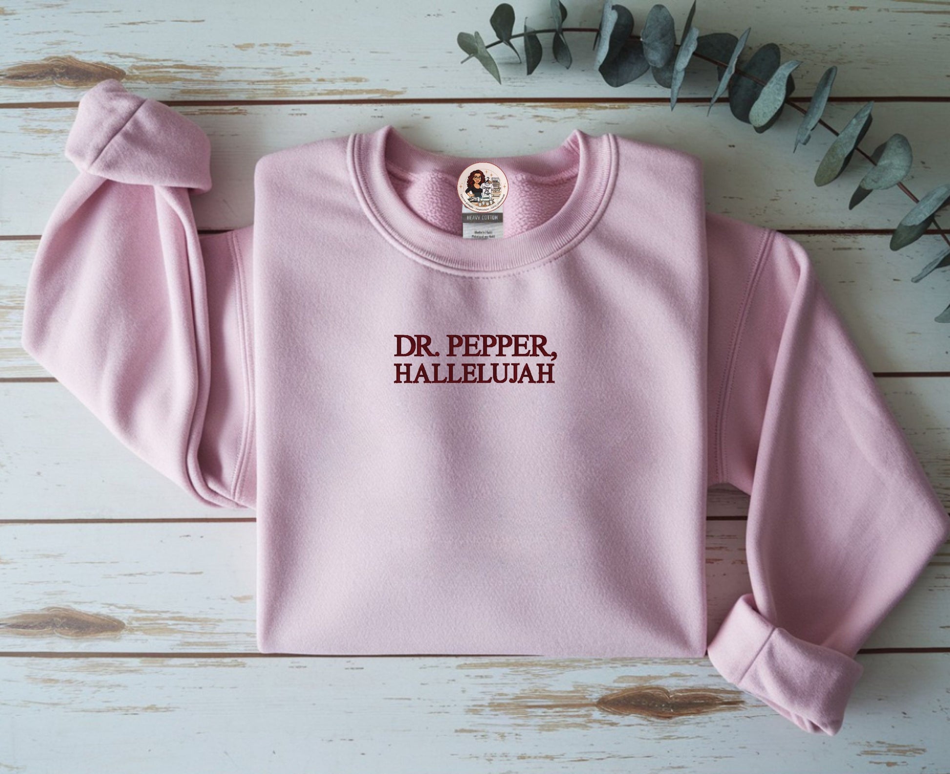 Pink sweatshirt with 'DR. PEPPER, HALLELUJAH' text on a wooden surface with eucalyptus leaves.