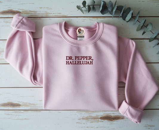 Pink sweatshirt with 'DR. PEPPER, HALLELUJAH' text on a wooden surface with eucalyptus leaves.