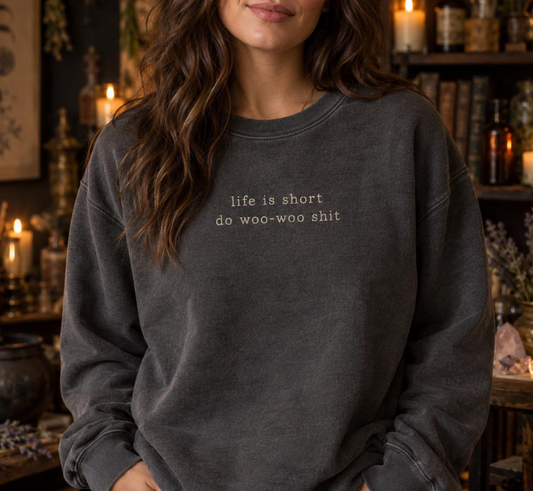 A woman wearing a dark gray crewneck sweatshirt with the text 'life is short do woo-woo shit' printed in white on the chest, set against a dimly lit, atmospheric background with candles and books.