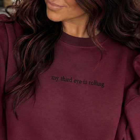 A woman wearing a burgundy crewneck sweatshirt with the text 'my third eye is rolling' printed in black on the chest, shown in a close-up lifestyle shot.