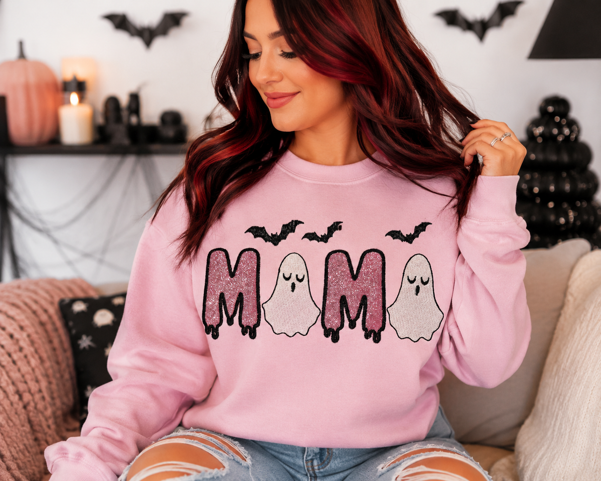 A woman with red hair wearing a pink crewneck sweatshirt with a 'MAMA' Halloween graphic featuring bats and ghosts, sitting on a couch in a decorated living room.