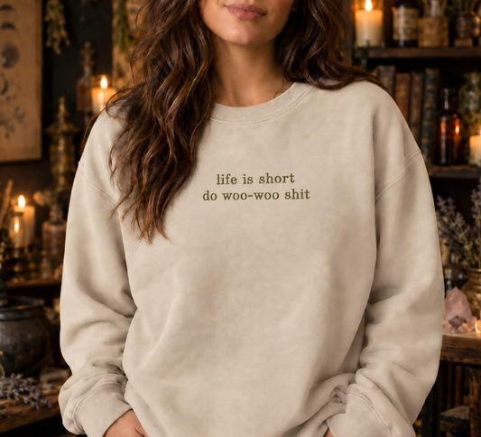 A woman wearing a cream-colored crewneck sweatshirt with the text 'life is short do woo-woo shit' printed in black on the chest, set against a dark, atmospheric background with candles and books.